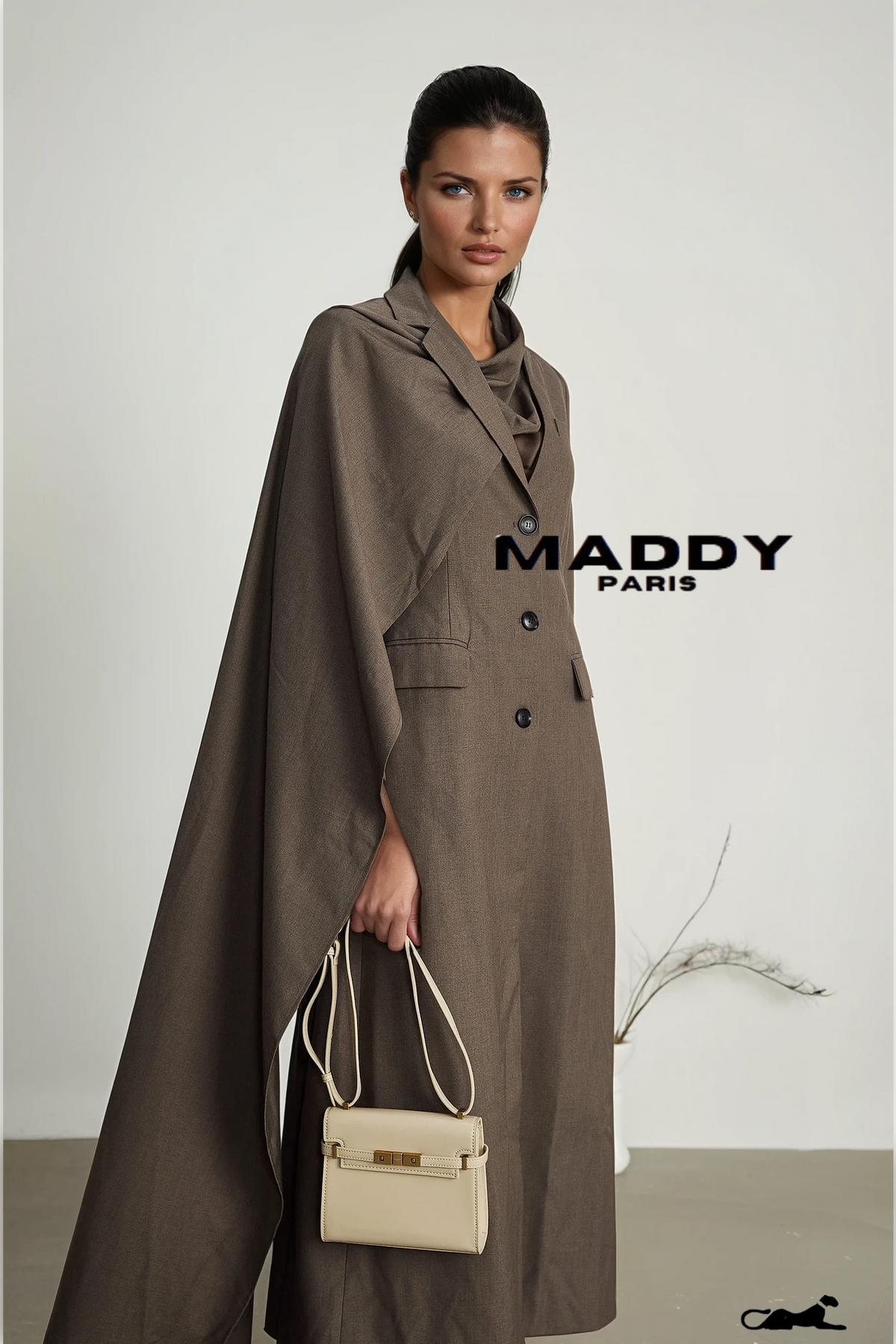 Woman wearing a long brown coat with a beige handbag, standing against a plain background with 'MADDY PARIS' branding.
